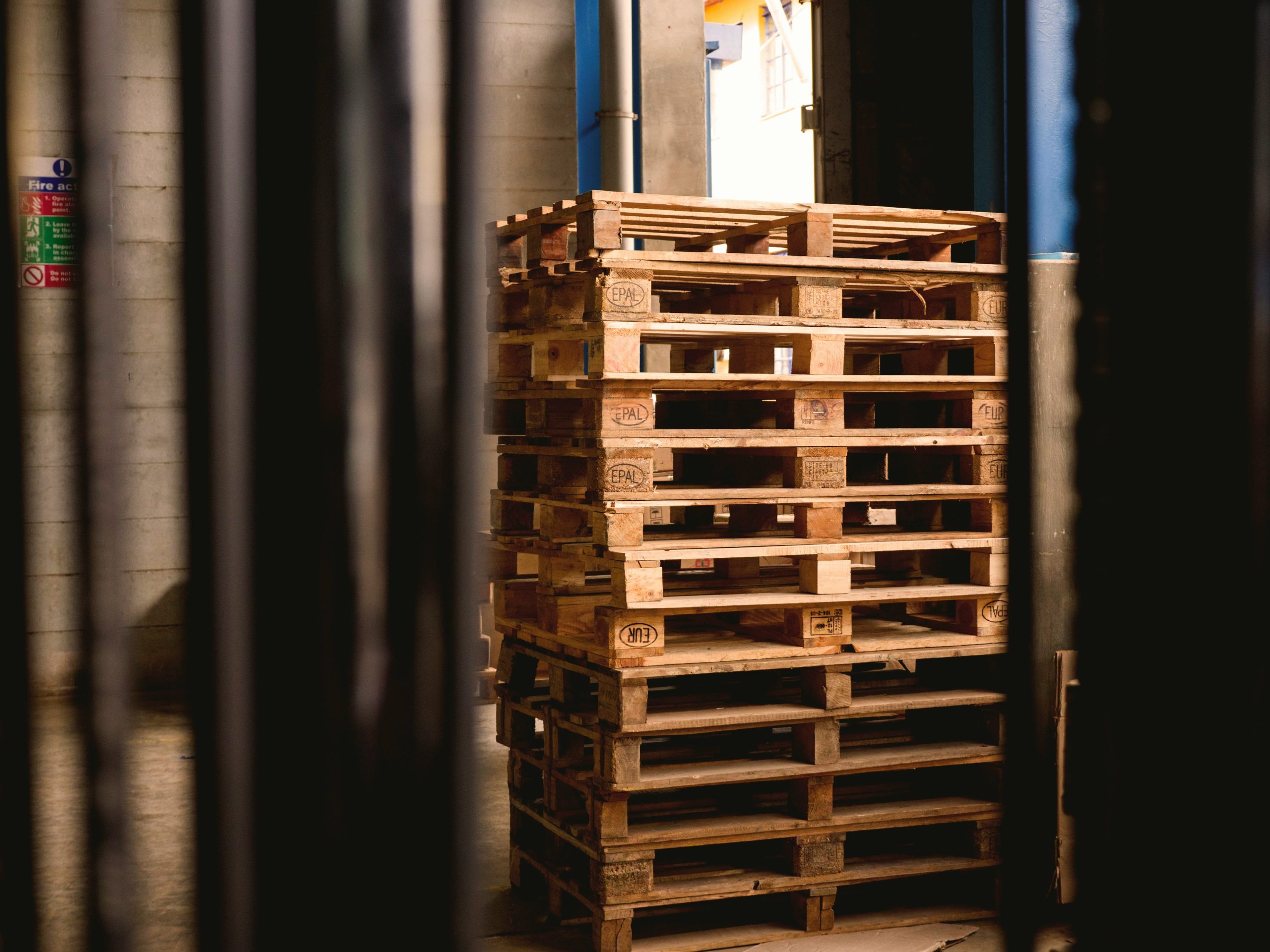 Stivă de paleți din lemn, marcați EPAL, depozitați într-un spațiu industrial interior, surprinși printre structuri metalice, cu lumină naturală care pătrunde din exterior. Contact Concept Pal Business
